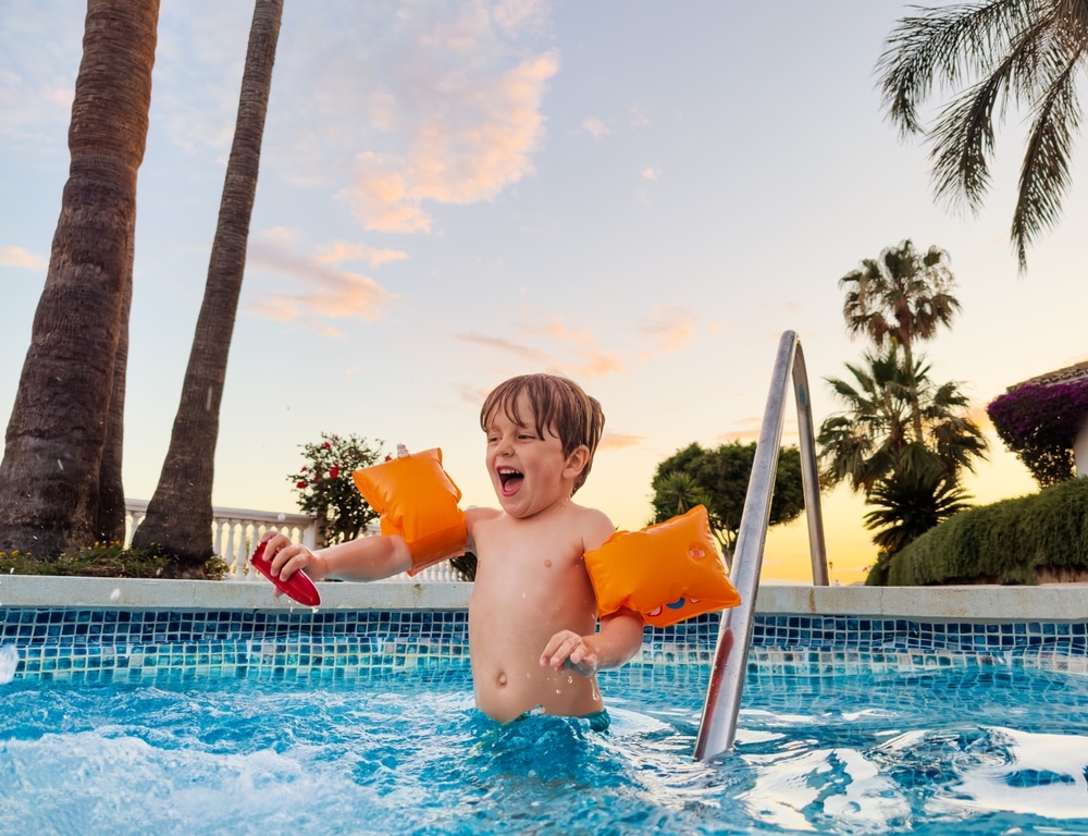 Joyful boy in arm bands enjoying sunset swimming pool having fun laughing and splashing 