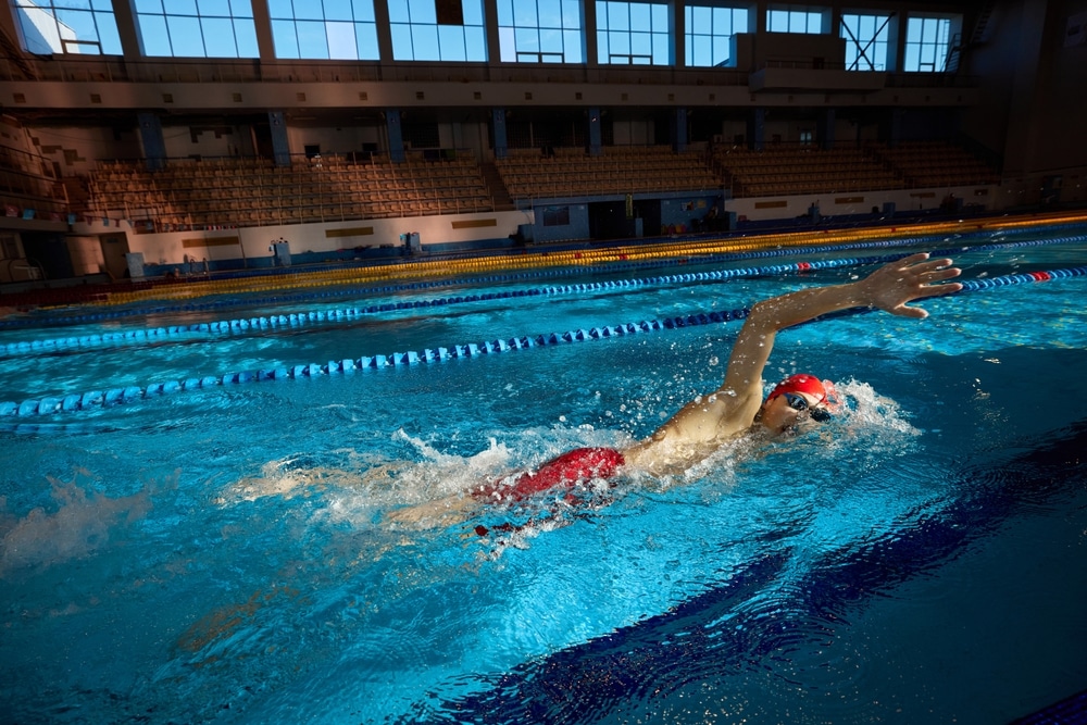 Young man, swimmer in red cap and goggles in motion, training freestyle stroke, swimming in pool indoors